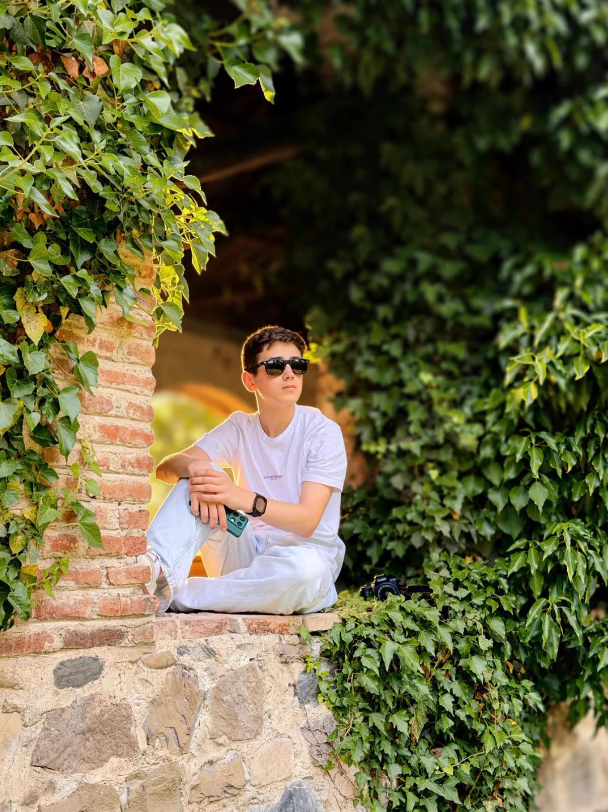 Sitting on a stone ledge surrounded by green ivy, wearing sunglasses
