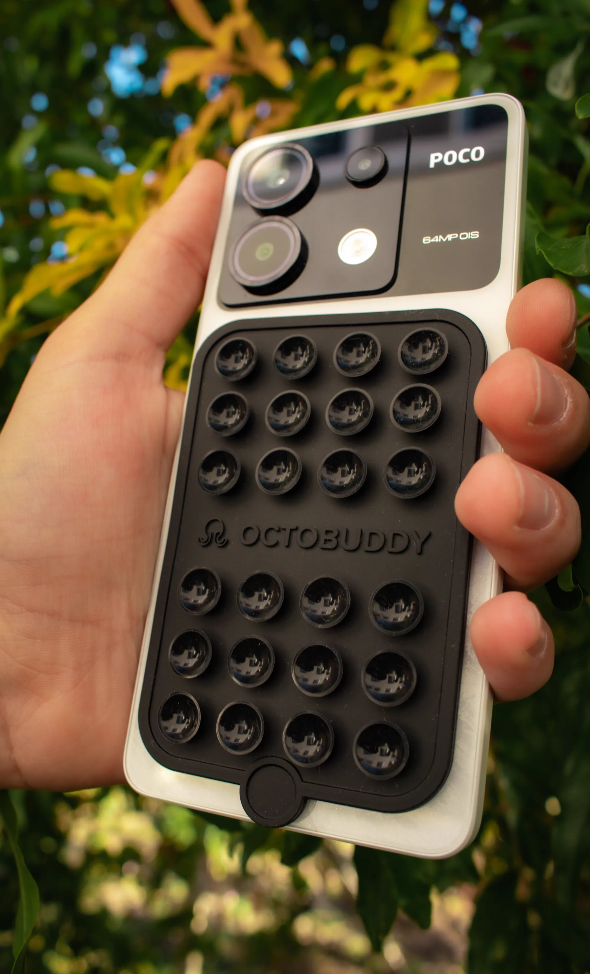 A hand holding a smartphone with an accessory on its back, surrounded by greenery