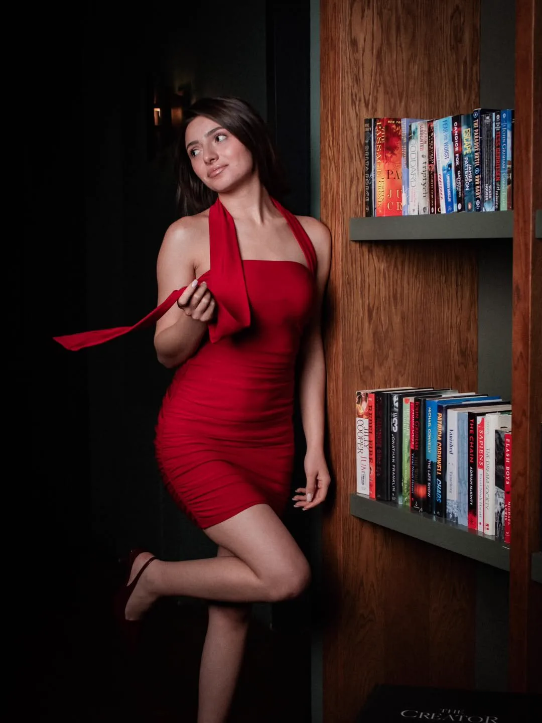 A woman in a red dress holding a red scarf next to a bookshelf